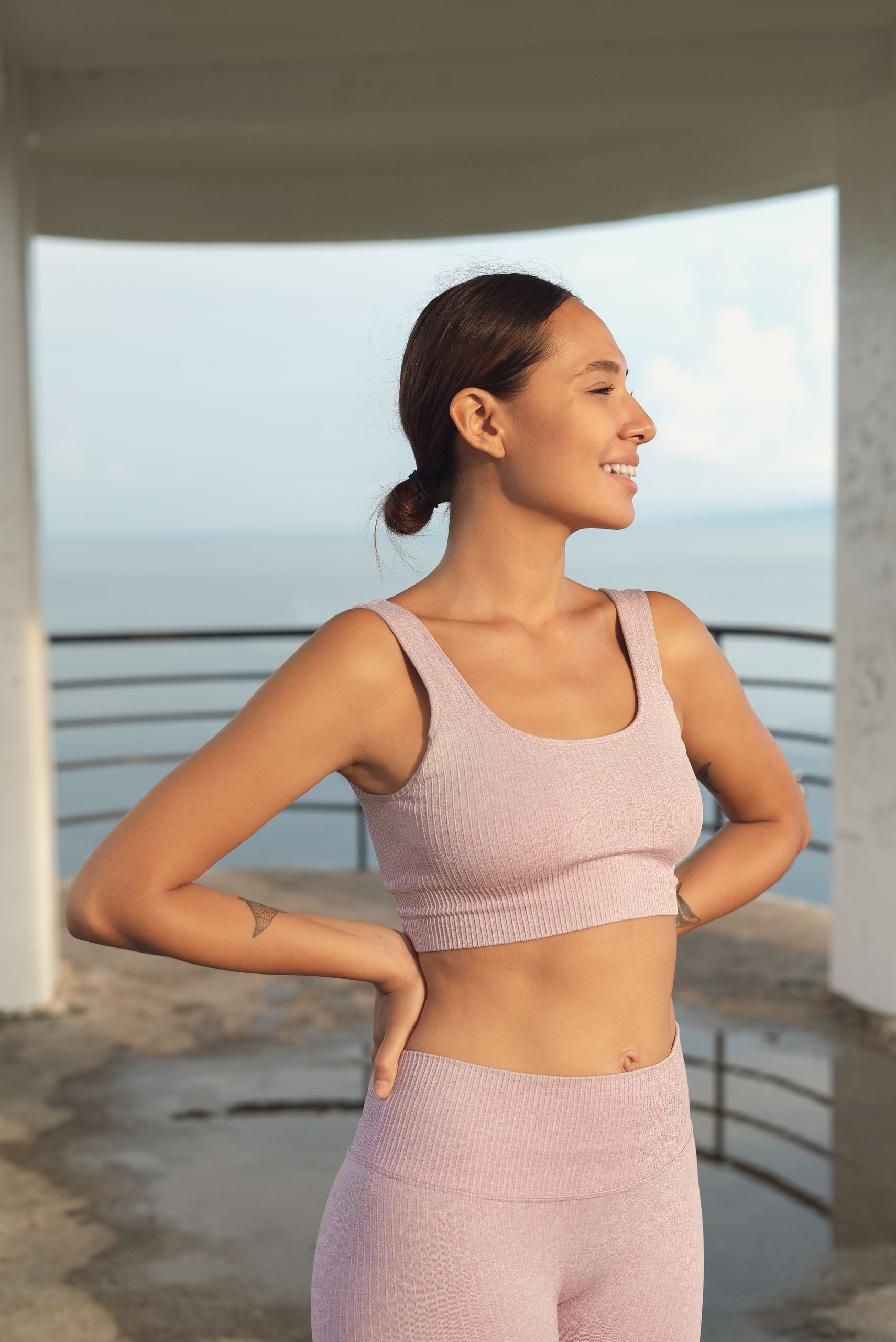 Confident woman in athletic wear standing by the sea, smiling in the sunlight.